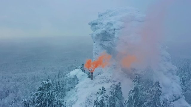 Aerial view: person holding orange signal smoke stands on top of a mountain surrounded by frosted snow covered forest. Female waving hands with signal flare, call for help, SOS, snowfall, blizzard.