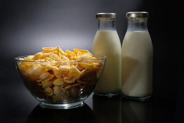 A bowl of cornflakes and milk in a glass on a dark background.