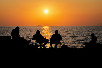 Fototapeta na wymiar Silhouettes of a group of happy friends resting after a hard day at the beach on beautiful sunset. People sitting and talking to each other