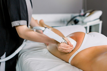 Female doctor cosmetologist doing roller massage with apparatus on female client thighs, close up. Roller massage in medical spa center