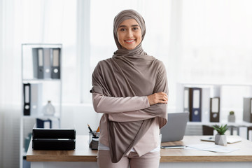 Confident Muslim Businesswoman Standing With Folded Arms Near Workplace In Office