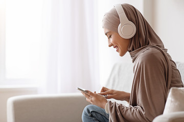 Muslim girl using smartphone for listening music in headphones at home