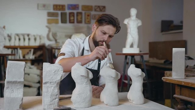 Attractive portrait of serious skilled pleasant bearded sculptor which carefully cleaning down the remnants of limestone from handmade sculptures