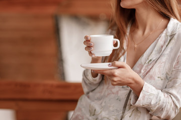 Young woman on the balcony holding a cup of coffee or tea in the morning. She in hotel room looking at the nature in sumer. Girl is dressed in stylish nightwear. Relax time