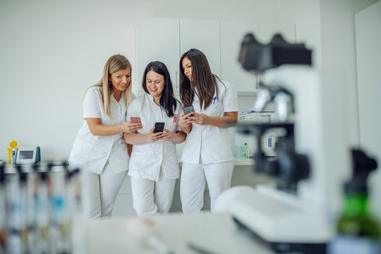 Three smiling cheerful Caucasian female lab assistants in white uniforms standing in lab and using smart phones.