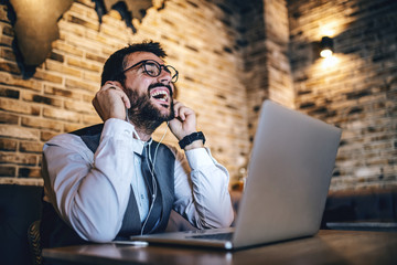 Attractive smiling caucasian bearded elegant businessman in suit listening music over laptop while sitting in cafe.