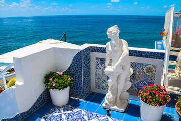Un precioso balcón con macetas, azulejos azules, y una escultura blanca con vistas al mar