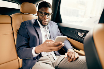 Businessman in a suit sits in a car back seat using his phone.