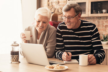 Cheerful senior couple watching news on laptop online at kitchen