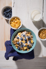 Bowl with oatmeal, corn flakes, blueberries and a glass of milk on a white wooden background