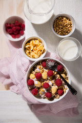 Bowl with oatmeal, corn flakes, raspberry and a glass of milk on a white wooden background