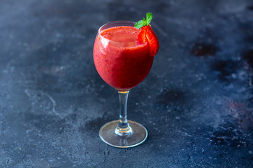 Fresh iced strawberry smoothie in wine glass decorated with slice of strawberry and mint on a dark background. Summer Slushy drink. Close up, copy space for text.