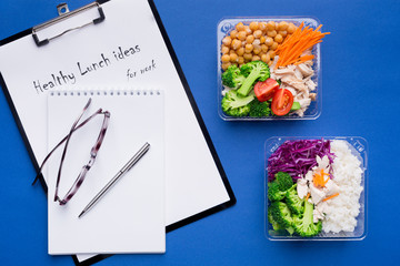 Lunch box with healthy food tablet, notebook for notes, pen and glasses on a bright blue paper background. The concept of healthy eating while working in the office. Top view