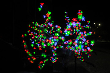 Colorful light bulbs in the form of flowers. A shining tree of multicolored flowers. A bright garland, light bulbs. Festive evening illumination, flickering lights, illumination.