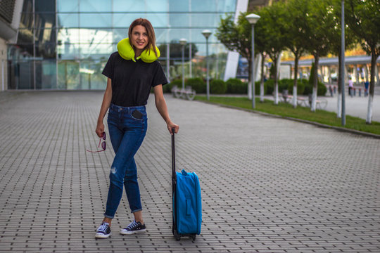 Full height portrait of young beautiful woman in blue jeans and black t-shirt with blue suitcase and yellow travel pillow. Early in the morning at the entrance to the airport terminal, natural light.