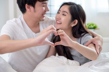 Obraz na płótnie Canvas Close up young Asian couple doing heart gesture together by hands and smiling on white bed in bed room