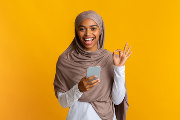 Black muslim girl in hijab using smartphone and showing ok gesture