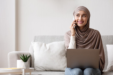 Smiling muslim woman talking on cellphone while using laptop at home