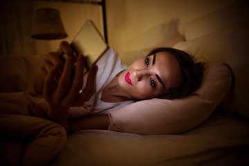 Staying up late. Close-up photo of a beautiful girl, who is wide-awake at night, lying in her bed and reading something from the screen of a smartphone.