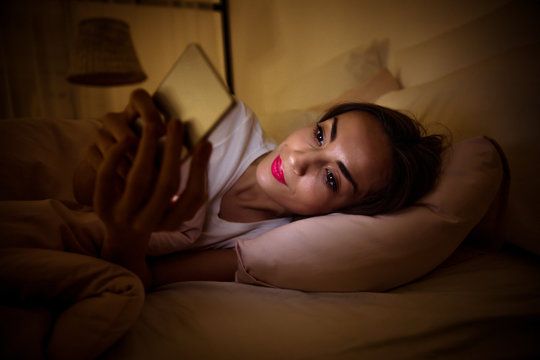 Staying up late. Close-up photo of a beautiful girl, who is wide-awake at night, lying in her bed and reading something from the screen of a smartphone.