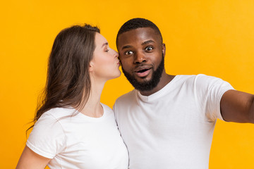 Obraz premium Young interracial couple in love taking selfie together, woman kissing boyfriend