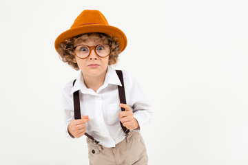 Beautiful boy with curly hair in white shirt, brown hat, glasses with black suspenders stands isolated on white background