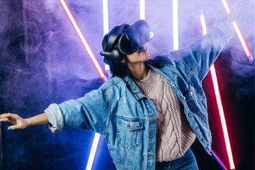 Mod curly dark haired girl dressed in blue denim jacket uses the virtual reality glasses on her head in the dark studio with neon light and smoke fog