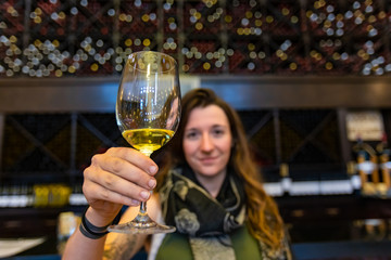 yellow white wine glass selective focus, caucasian young woman hold, drink and toasting the wineglass, tasting room wines display shelves background