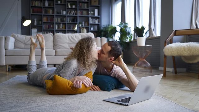 Couple watching computer in pajamas. Millennial Man and woman relaxing on carpet spending weekends morning together using laptop, watching sitcom or online shopping in modern European style flat 