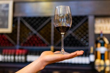 A glass of red wine on hand, selective focus close up view against tasting room bottles storage racks background, blurred copy space on sides