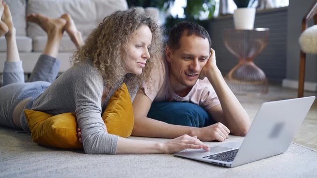 Couple watching computer in pajamas. Cropped portrait man and woman relaxing on carpet spending weekends morning together using laptop, browsing or online shopping in modern European flat 