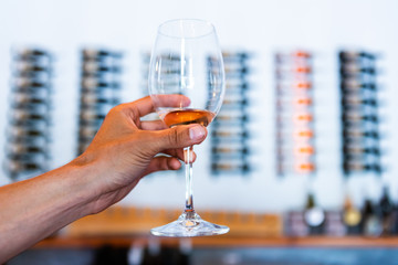 Man hand holding a glass of white wine in selective focus view, Wine bottles on wall storage racks and shelves, Wines stored blurred background