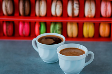 Assorted macaroon cakes in a red box and two white cups of coffee on a gray background.