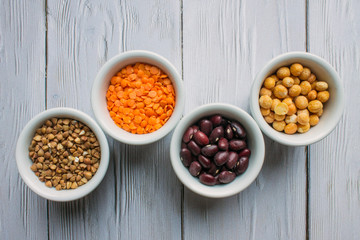 Four porcelain small cups with cereals top view: red beans, orange lentils, buckwheat and yellow peas on a white wooden background with space for copying