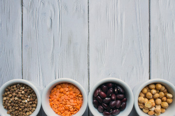 Four porcelain small cups with cereals top view: red beans, orange lentils, buckwheat and yellow peas on a white wooden background with space for copying