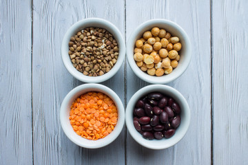 Four porcelain small cups with cereals top view: red beans, orange lentils, buckwheat and yellow peas on a white wooden background with space for copying