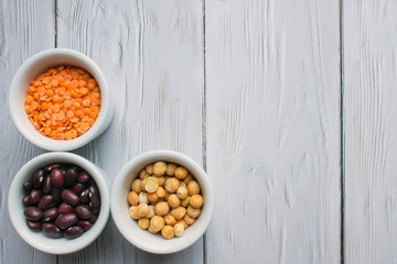 Three porcelain small cups with cereals top view: red beans, orange lentils and yellow peas on a white wooden background with space for copying