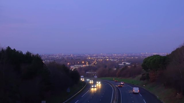 A highway full of moving cars towards a city lit by lights on the horizon