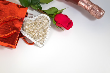 A red rose, a bottle of champagne and a heart symbol in a gift bag on a white surface on Valentine's Day. Copy space. Horizontal orientation.