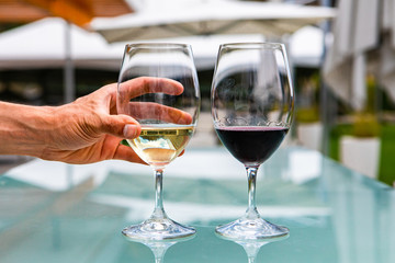 man hand on white wine glass, pair of white and red wines glasses close up selective focus, restaurant patio outdoor table background