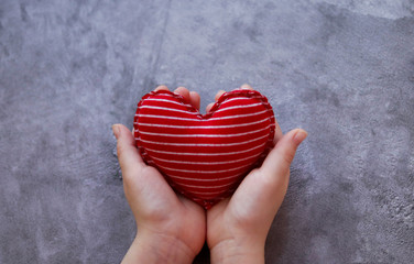 Textile soft red heart with white stripes in the hands on a gray concrete background.Selective focus.Cotton heart for Valentine's day, hand-sewn. A gift for February 14, made with your own hands