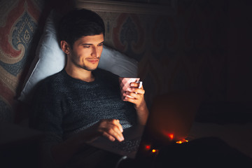 Night portrait of young cheerful man lying on bed in dark room at home with cup of coffee and laptop.