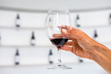 Man hand holding a glass of red wine, selective focus close up view against white tasting room with wine bottles wall rack shelves blurred background