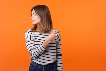 Get out! Portrait of resentful woman with brown hair in long sleeve shirt standing with closed eyes and offended face, pointing sideways showing exit. indoor studio shot isolated on orange background