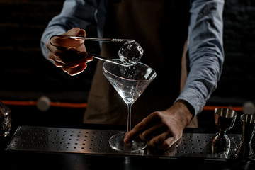 Fototapeta na wymiar Male bartender putting ice cube with twezzers to the tall martini glass