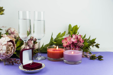 Fototapeta na wymiar Marriage proposal and love concept for Valentine`s day, box with wedding or engagement ring with brilliant. Glass of champagne, romantic dinner, peony flowers and candles on trendy purple background