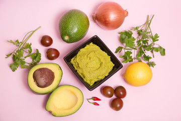 Guacamole en un bol con ingredientes sobre fondo rosa pastel.