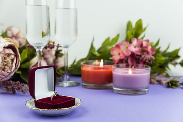Marriage proposal and love concept for Valentine`s day, box with wedding or engagement ring with brilliant. Glass of champagne, romantic dinner, peony flowers and candles on trendy purple background