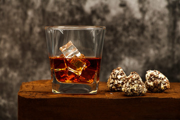 Whiskey with ice or brandy in a glass with truffles on a wooden table and a gray background. Whiskey with ice in a glass. Whiskey or cognac in a glass with truffles. Selective focus.