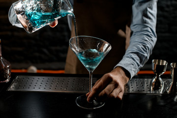 Professional male bartender pouring a blue alcoholic cocktail from the measuring cup with strainer to a martini glass